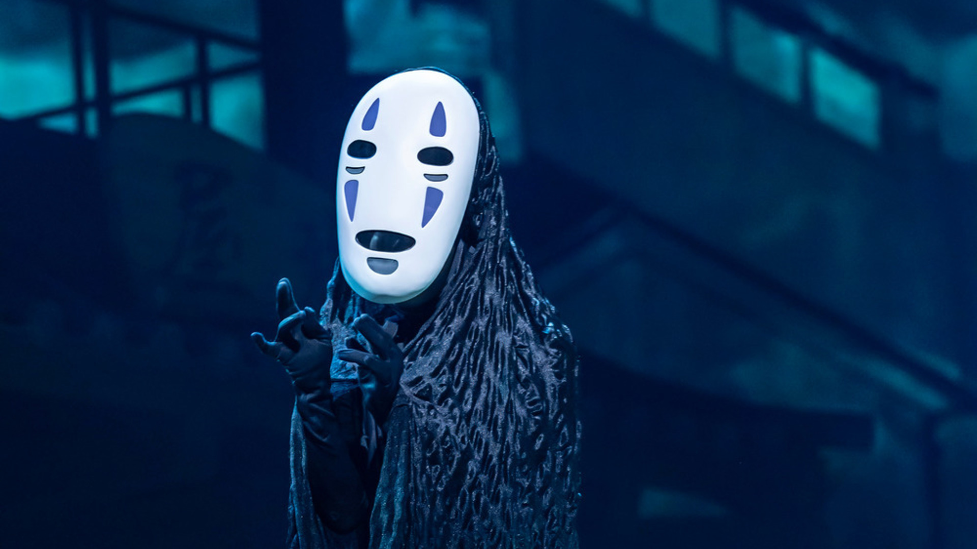 An actor stands on stage in a white mask, cloaked in black robes.