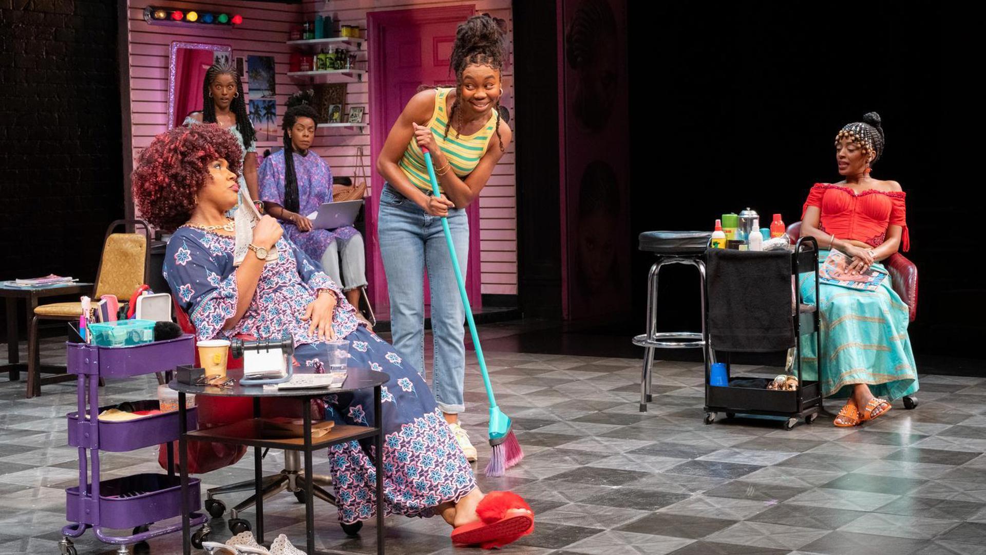 One performer stands with a broom, speaking to a performer who sits in a chair fanning themselves with a newspaper. Three performers observe from the background.
