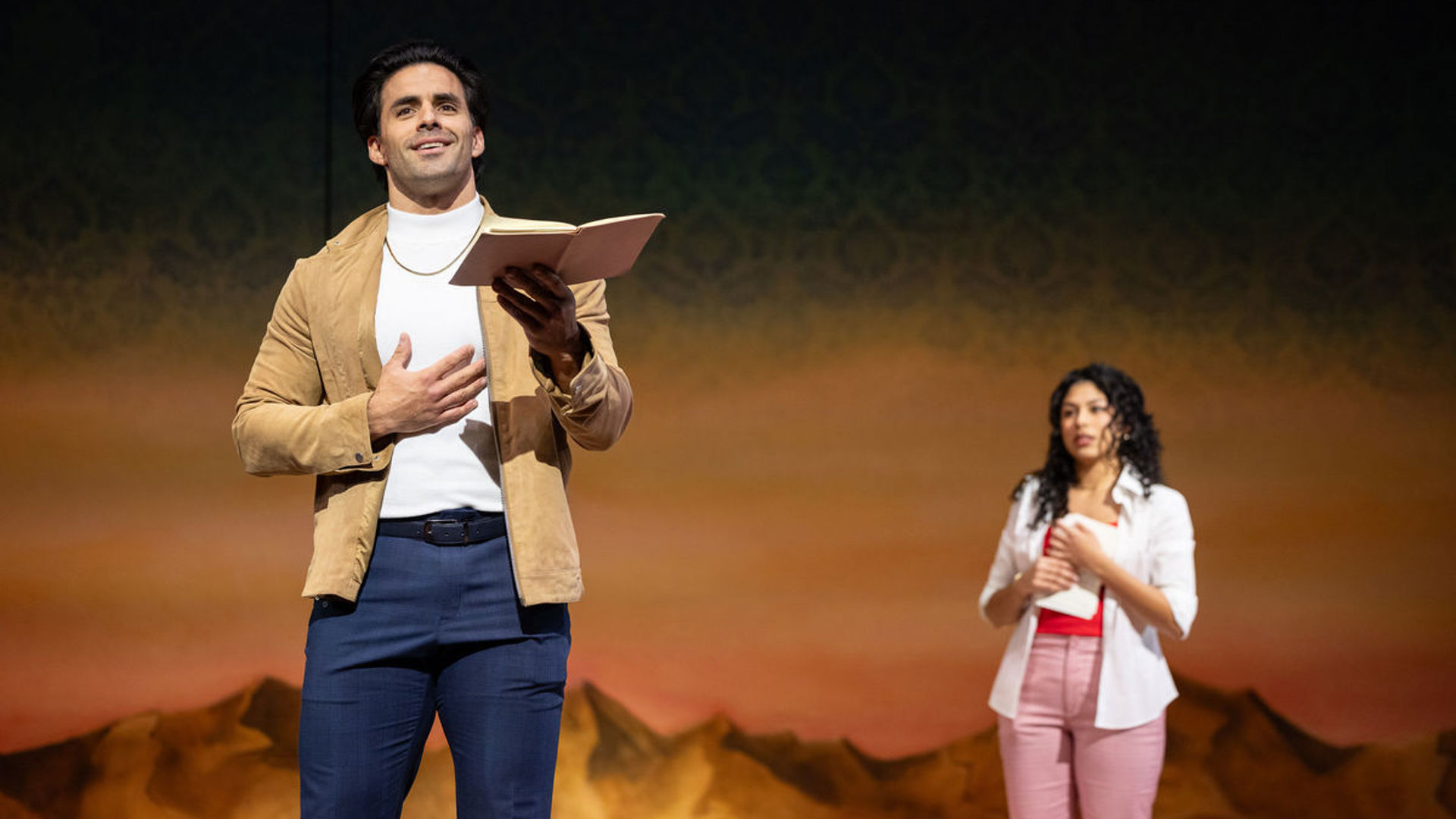 An actor stands downstage reading off of a book. An actor holding another book watches him from behind.