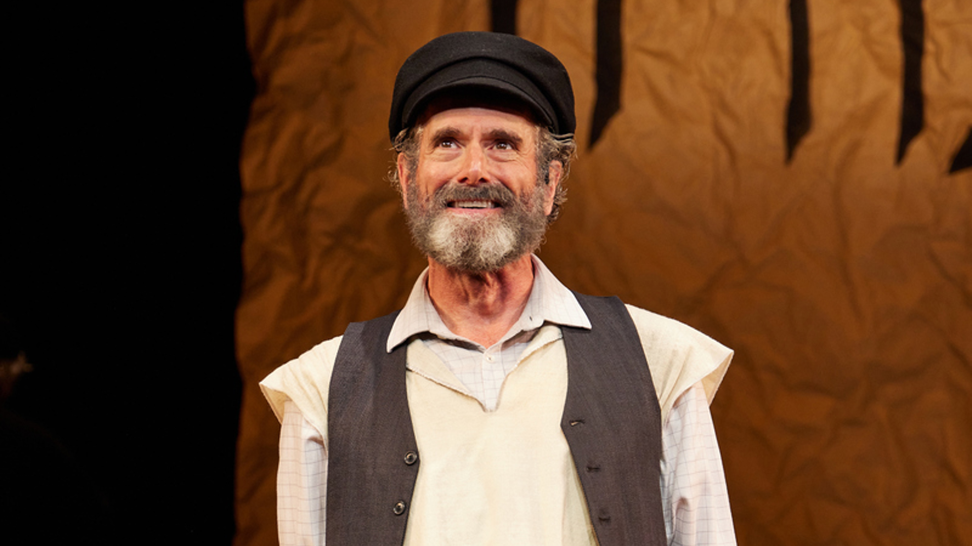 A man stands on stage stands smiling out at the audience. He is wearing a hat, a tallit and a vest. There is a wagon with a metal jug in it behind him.