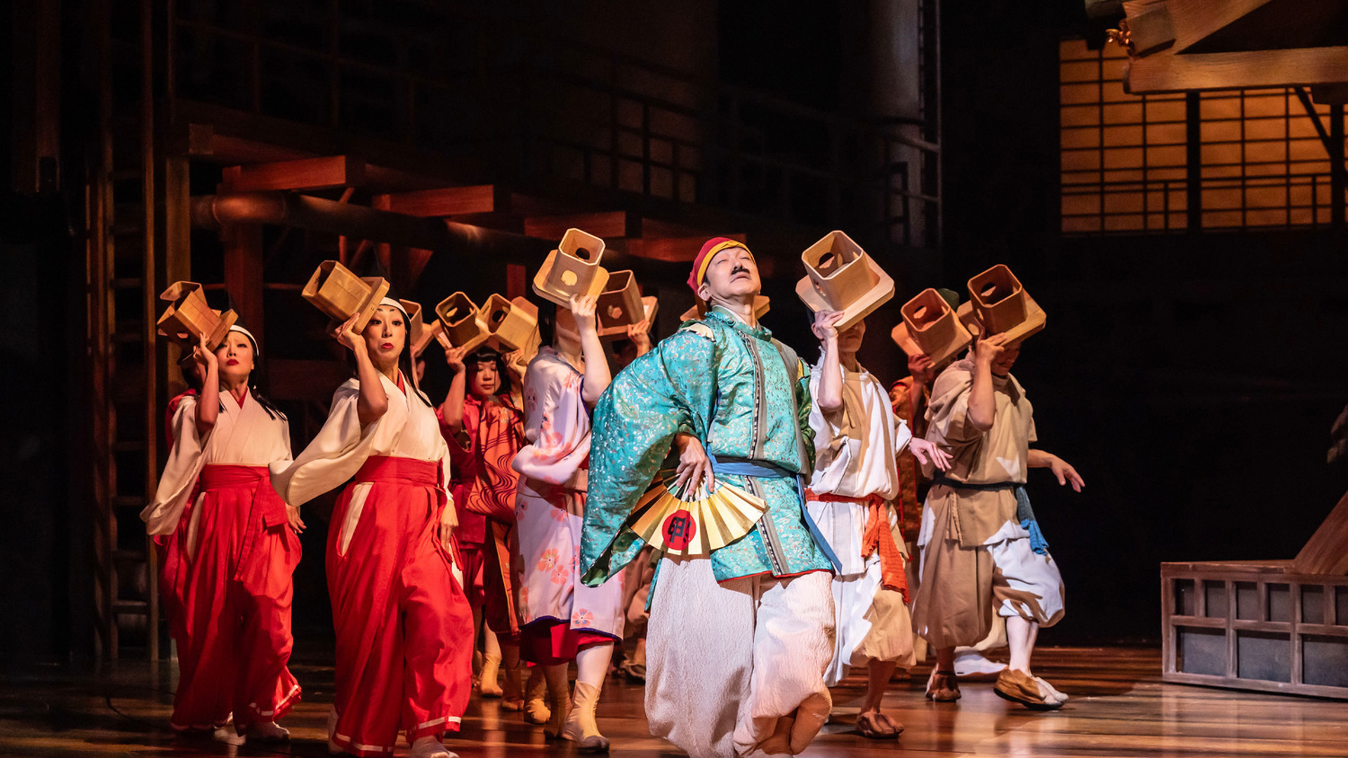 An actor holding a fan and wearing a blue robe steps towards the audience, leading a large group of actors behind him that are holding small, wooden boxes to their heads.