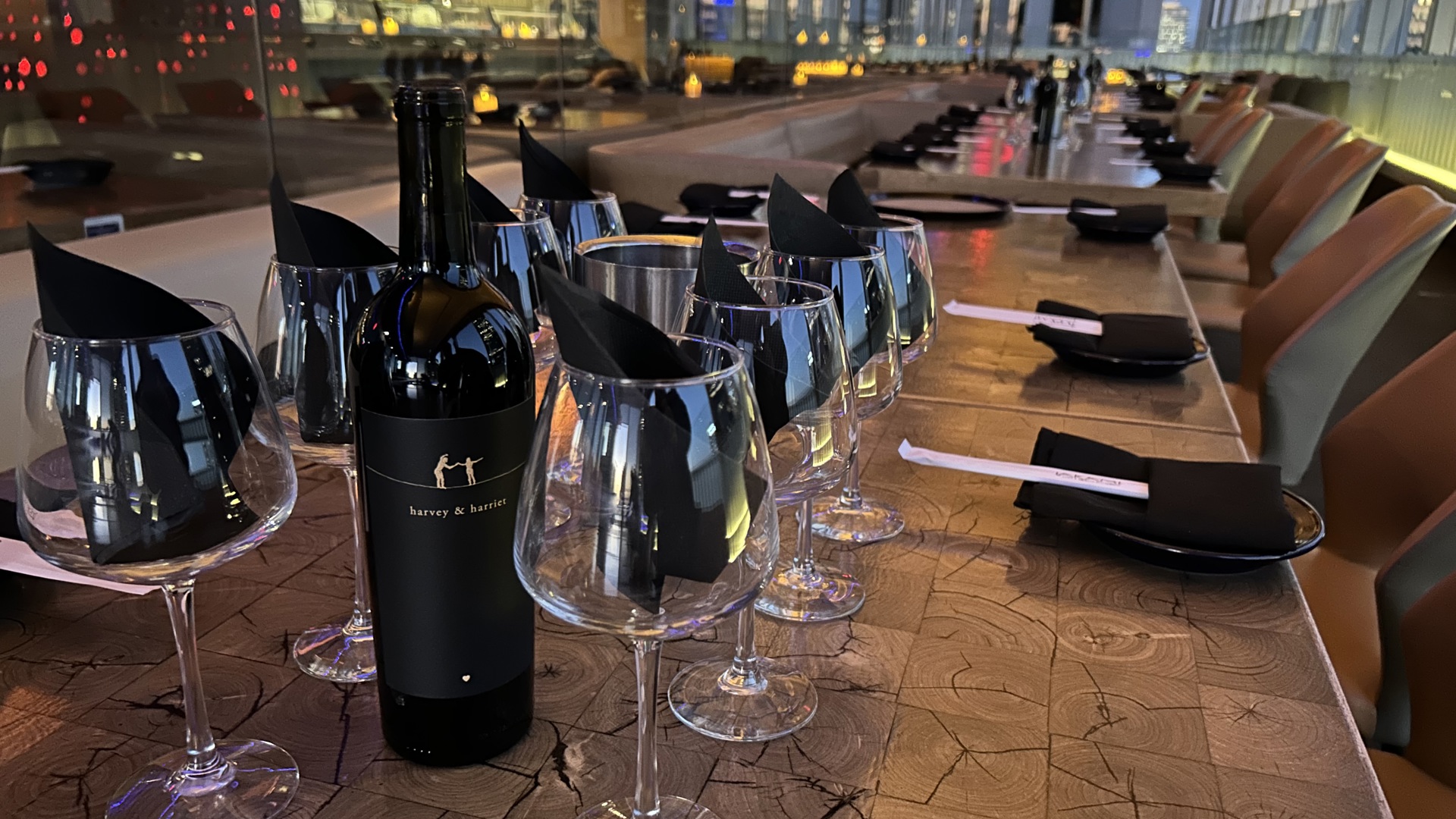 A bottle of wine sits on top of a wooden table with 8 neatly placed wine glasses with napkins inside them. 