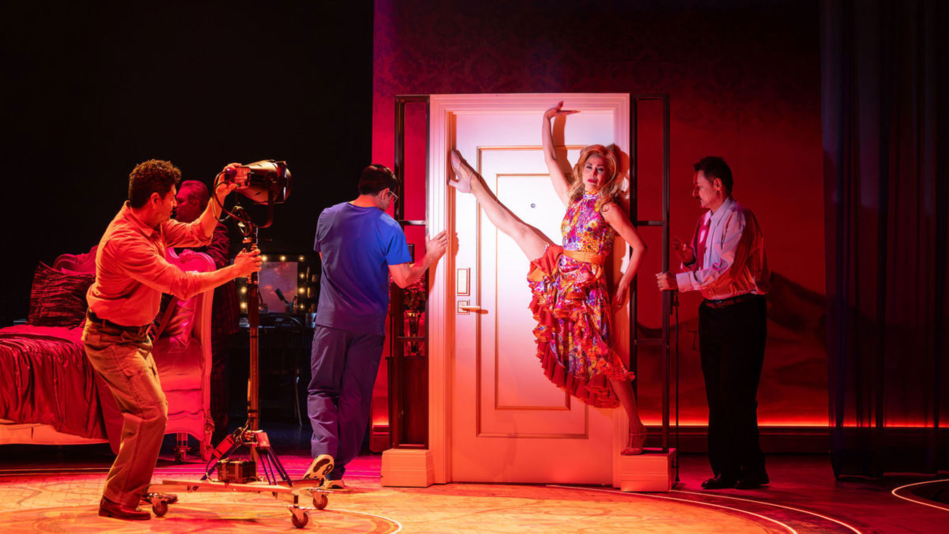 An actor points a spotlight at an actress stretching her leg against a door that being held by two other actors.