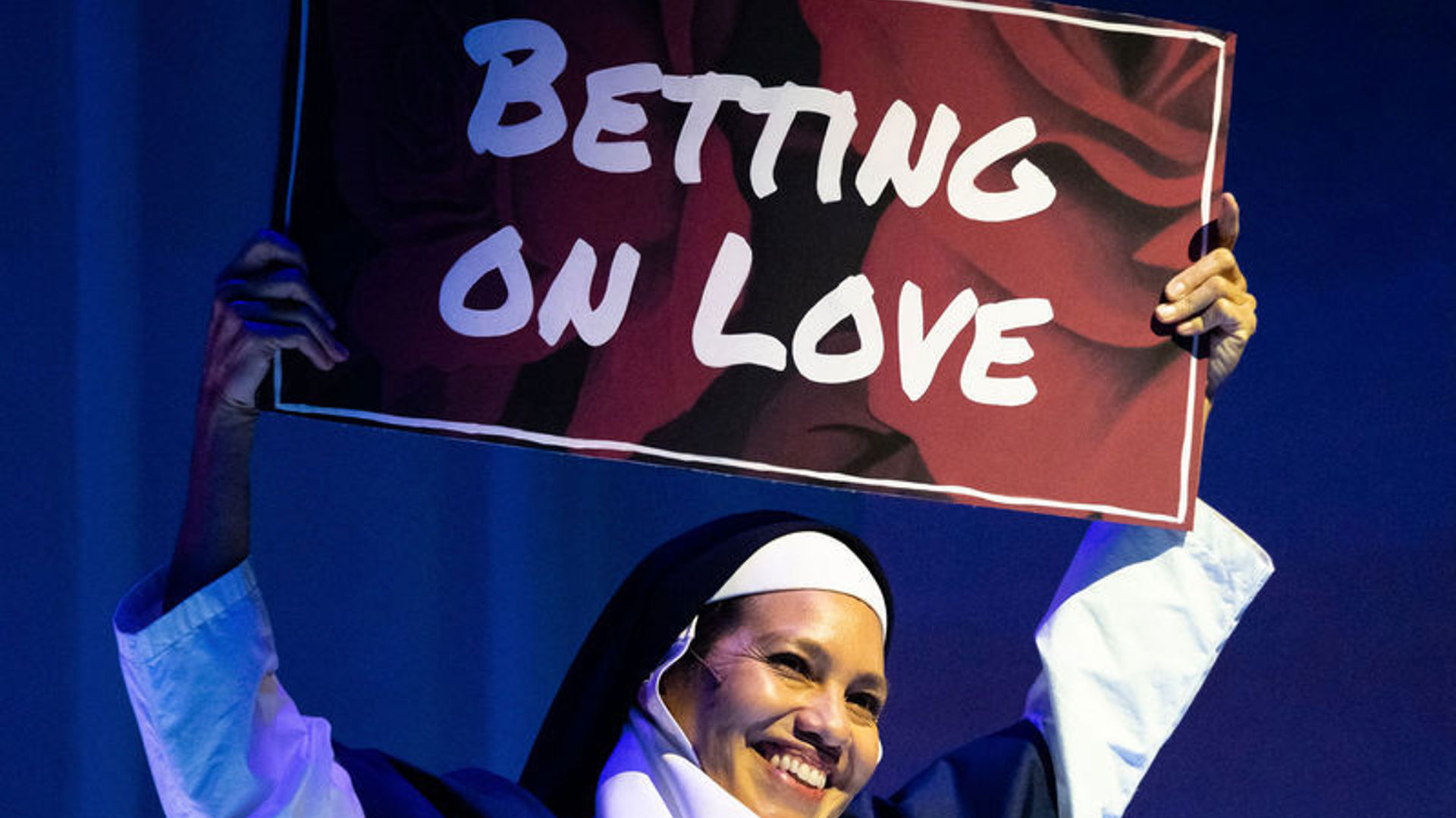 An actor dressed as a nun holds up a sign that reads 'BETTING ON LOVE'.