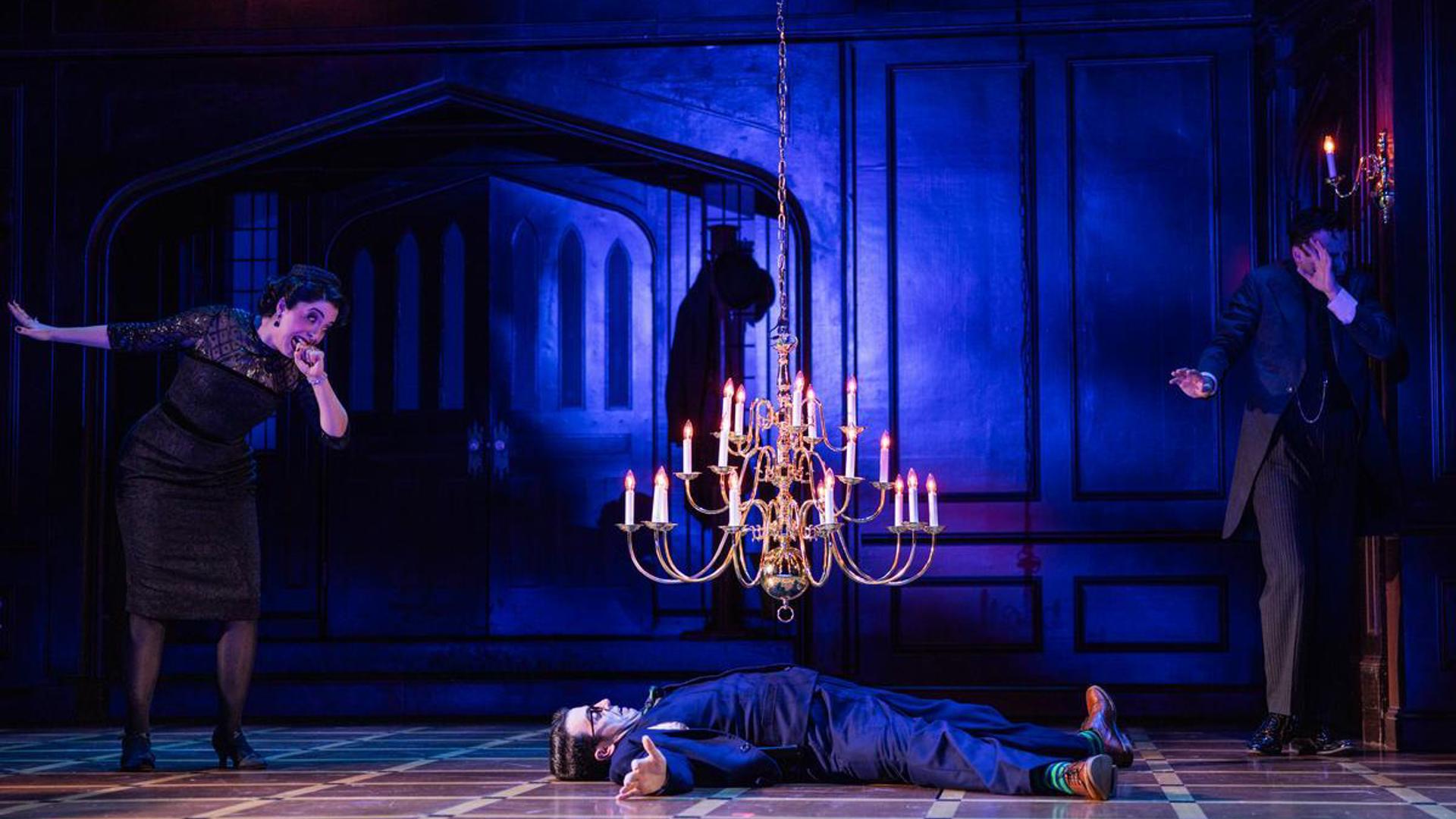 A man lays on the floor, mouth agape as a falling chandelier hangs over him. A woman stands to his left with her fist in her mouth and eyes wide watching the chandelier fall. A man stands to the right of the man on the floor, stretching his arms out and looking away from the chandelier.