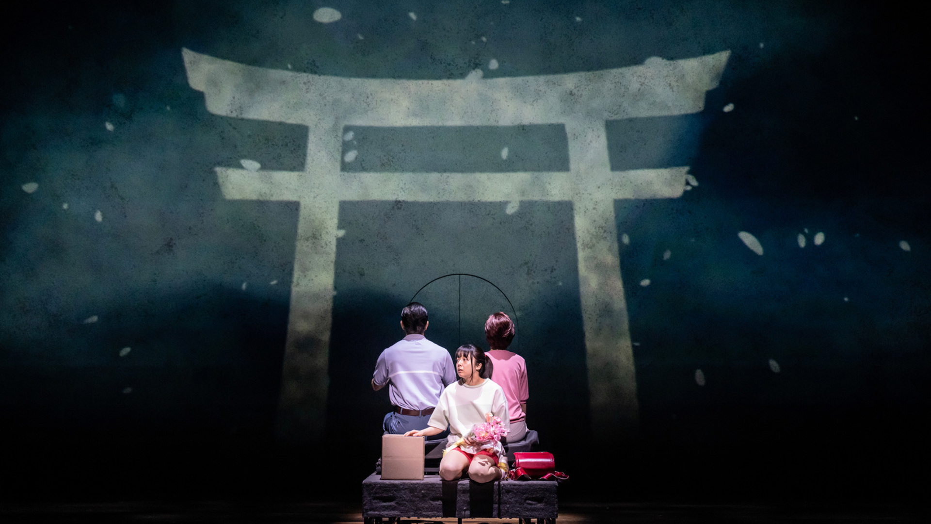 An actor sits on the back of a car, looking at the audience and carrying a bouquet of pink flowers. Her parents sit behind her, facing a gate projected on the back of the stage.