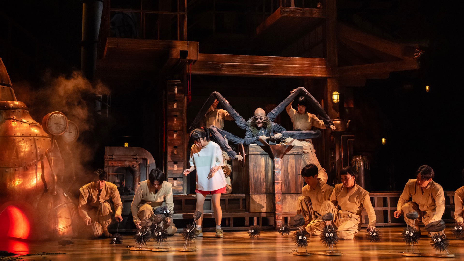 An actor in the center of the stage controls many switches with four arms. Another actor stands in front of him, looking at multiple small black puff creatures with eyes that are carrying coal.