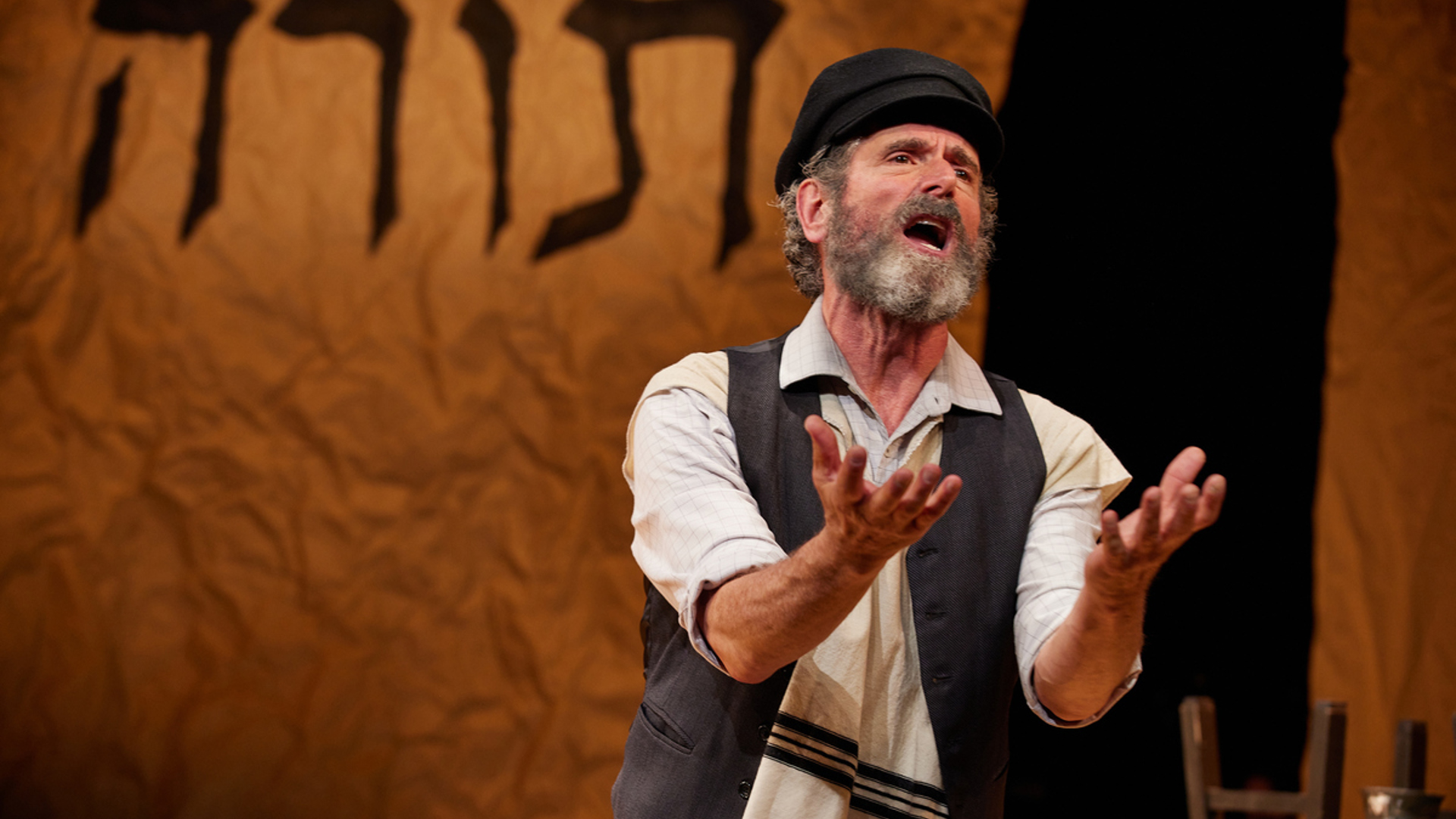 A man gestures towards the audience with his arms out as he sings. He is wearing a black hat, a gray vest and a tallit.