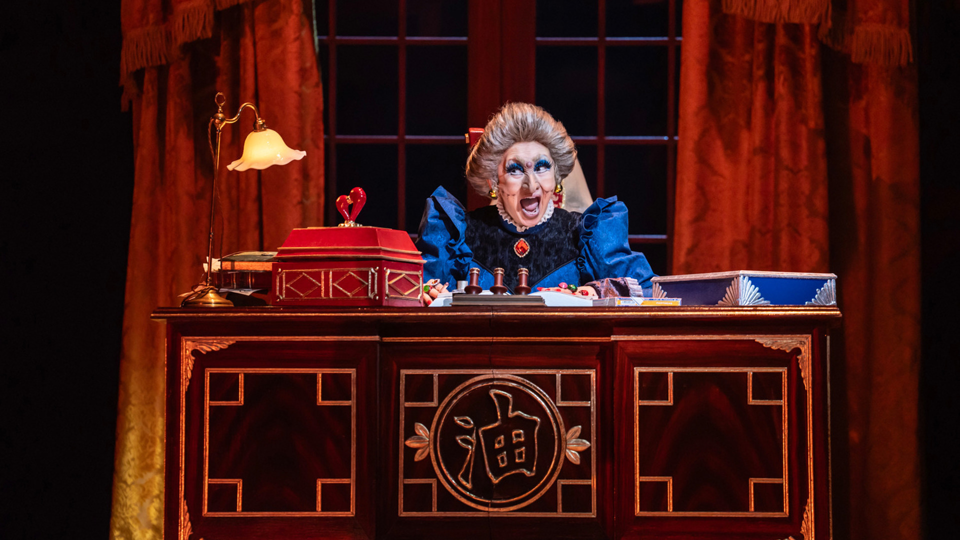 An actor sits behind a large wooden desk covered in writing materials. There is a window and grand red curtains behind her.