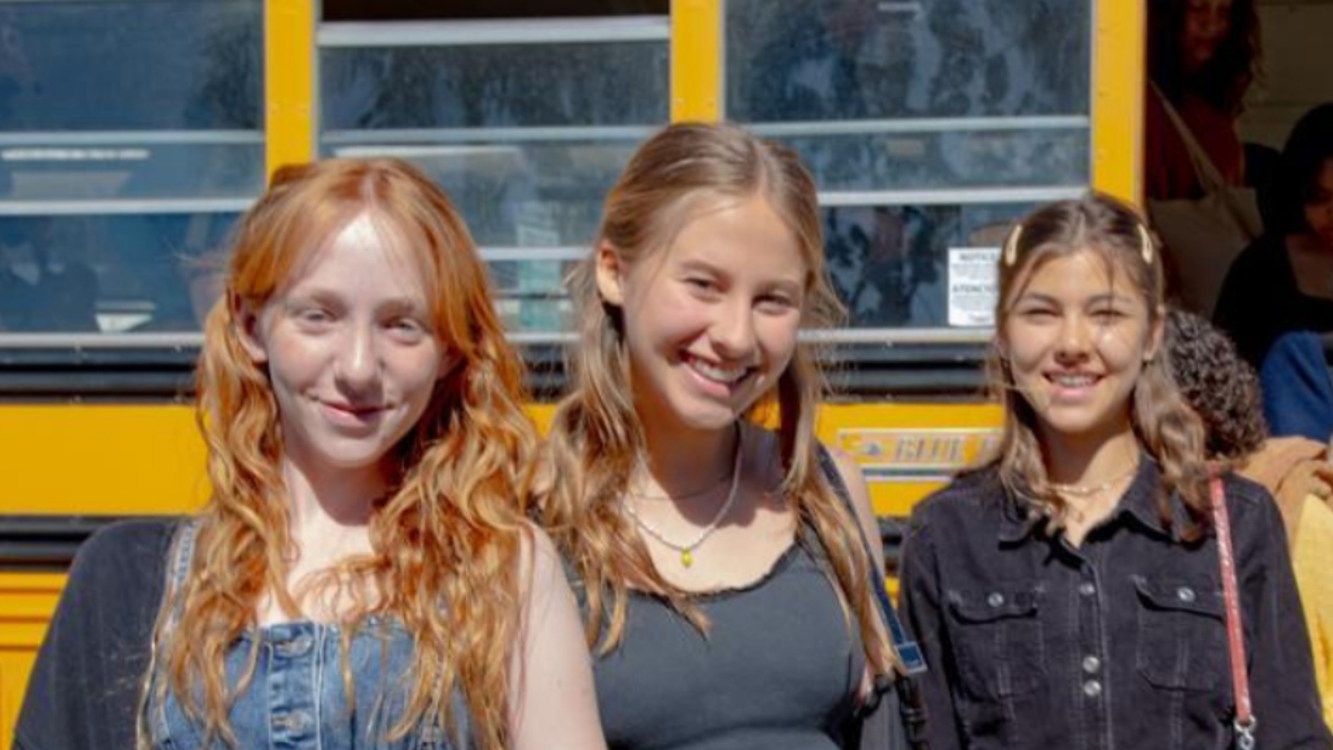 Three smiling teenage girls standing in front of a yellow school bus as other students step off behind them.