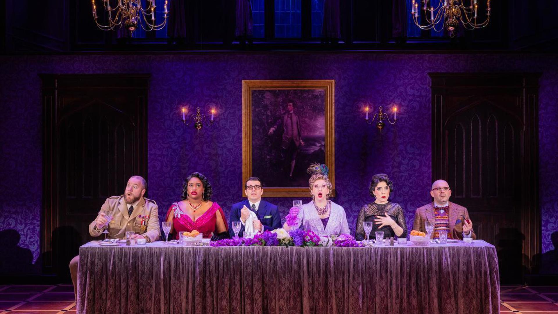 Six actors in formal attire sit all on the same side behind a long fancy dinner table. Each actor is staring forward into the distance with a shocked and perplexed look.