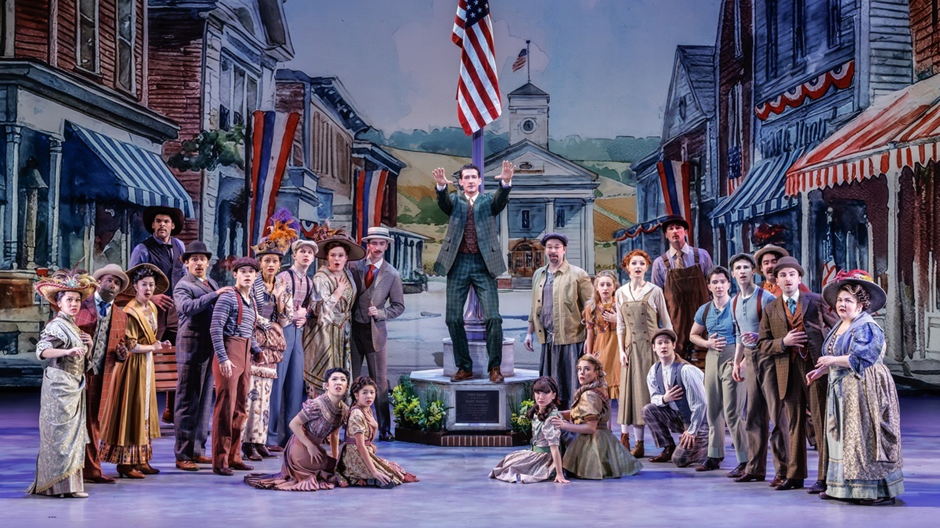 A large company of actors stand on stage in a semicircle looking out at the audience, one actor stands in the center with both arms extended to the audience on an elevated pedestal containing a flag pole with American flag. There is a backdrop behind them depicting the main street of a town.