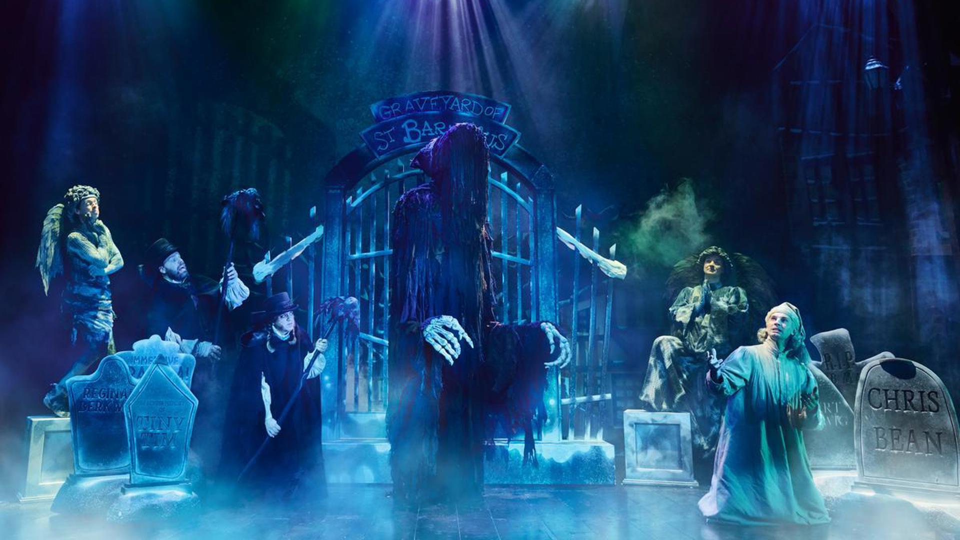 There is a large cloaked puppet in the middle of the stage, with 5 actors standing around it amongst gravestones.