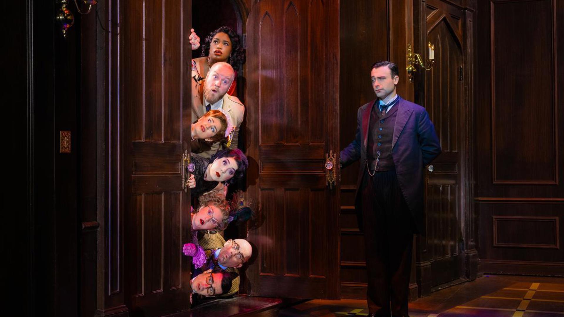 Seven actors lay stacked on top of each other horizontally, peeking out from behind an open door. Only their surprised faces are visible outside of the door frame. Across from them, a man in a suit has one arm outstretched, having just opened the door.