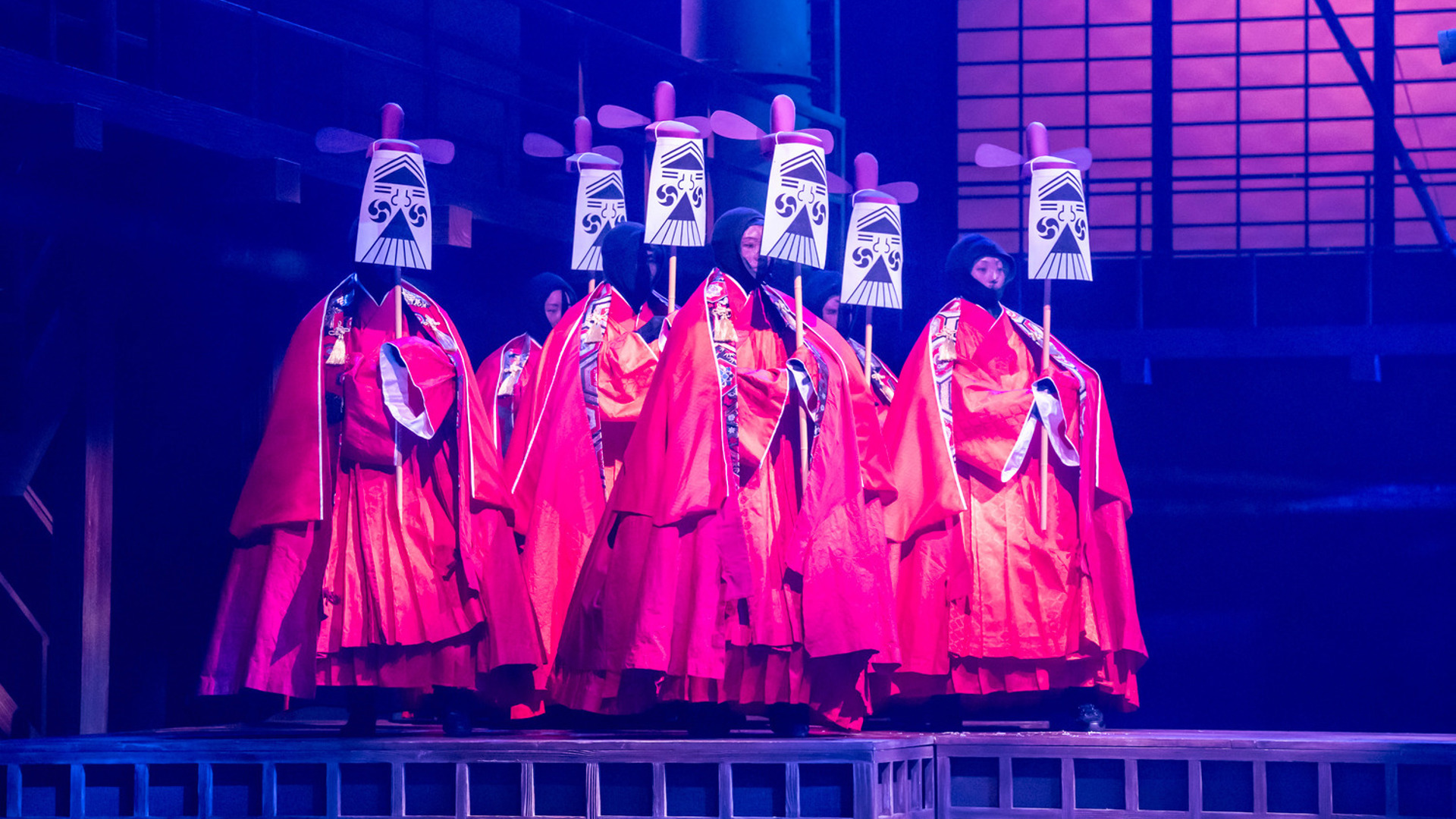 A group of actors in red robes march forward carrying masks on sticks.