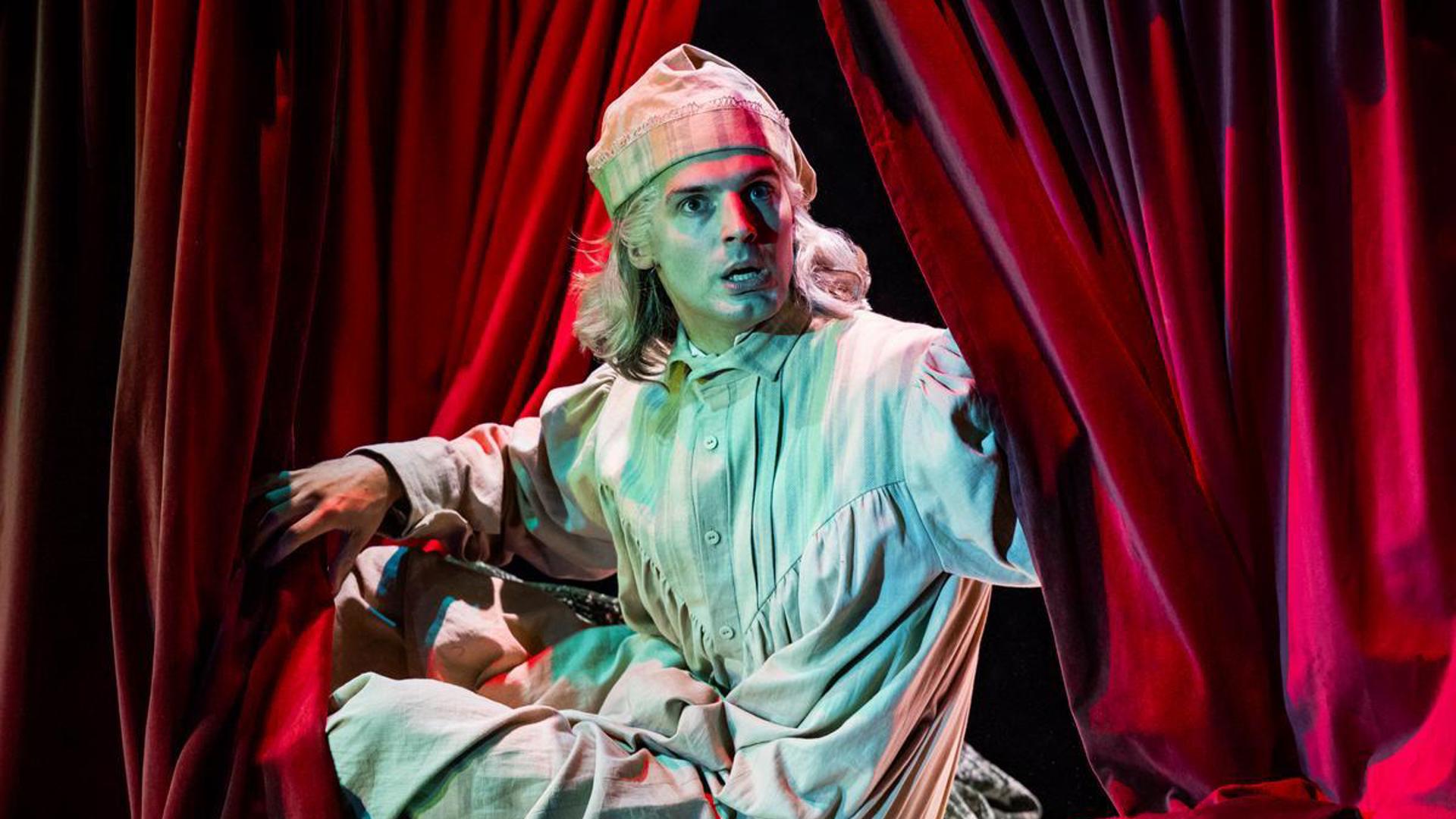 A performer sits up in bed and peeks through a curtain.