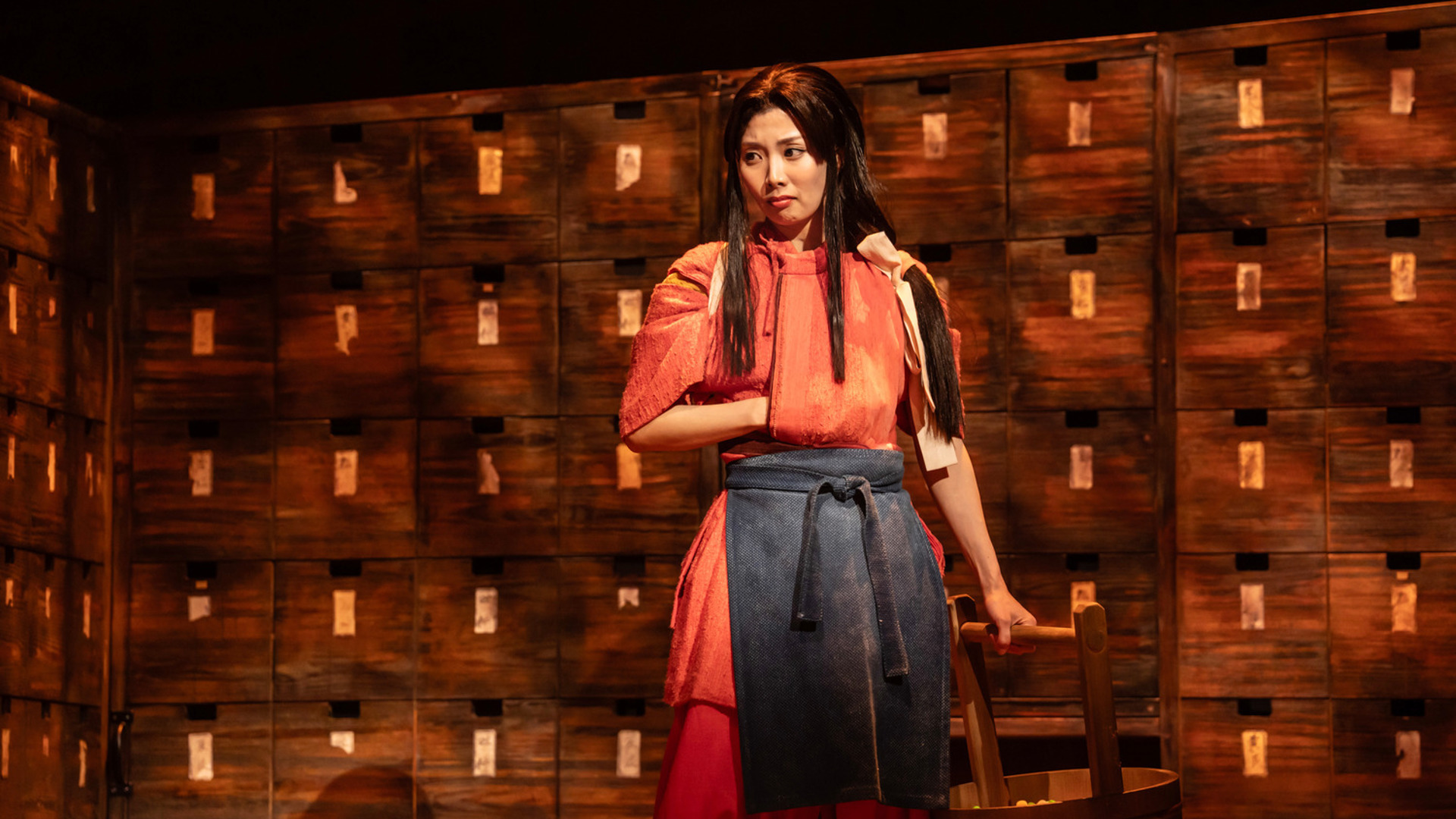 An actor stands onstage looking to her right. Her right hand is in her pocket and she has a bucket in her left hand.