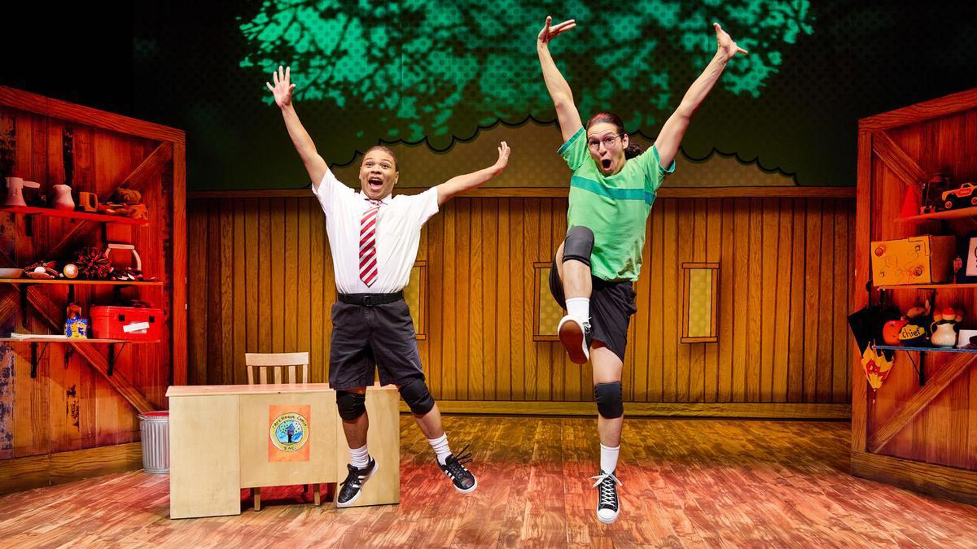 Two actors jump on stage with their arms and legs extended, behind them is a wooden desk and chair with shelves on the right and left adorned with many items.
