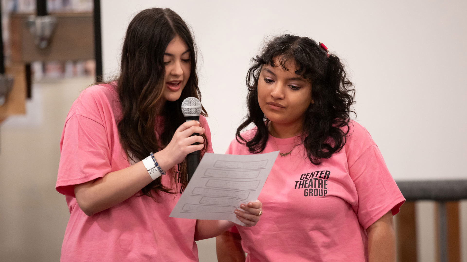 Two young women in pink Center Theatre Group T-shirts read from a paper, with one speaking into a microphone.