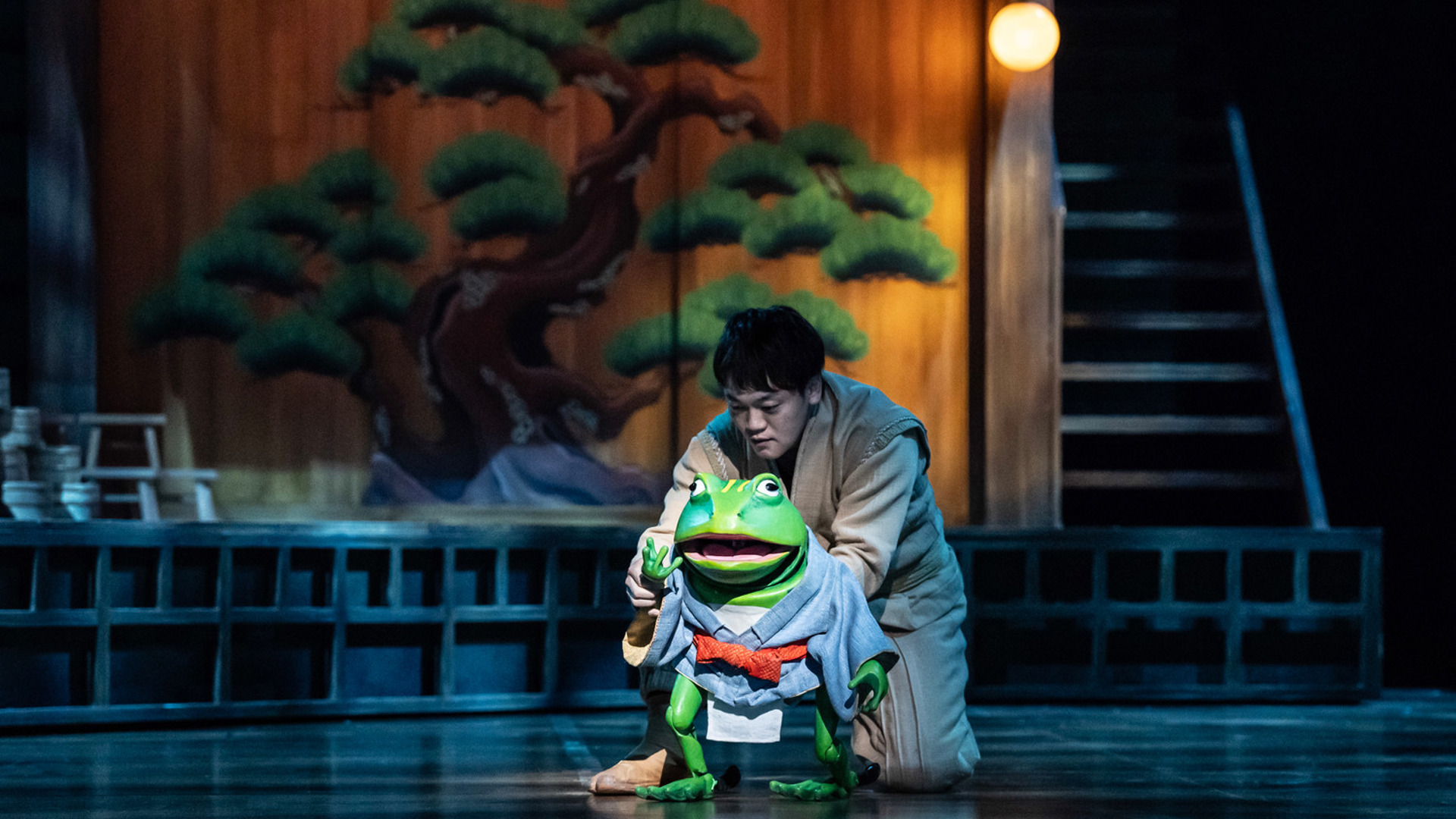 An actor crouches behind a frog puppet which is wearing a blue robe.