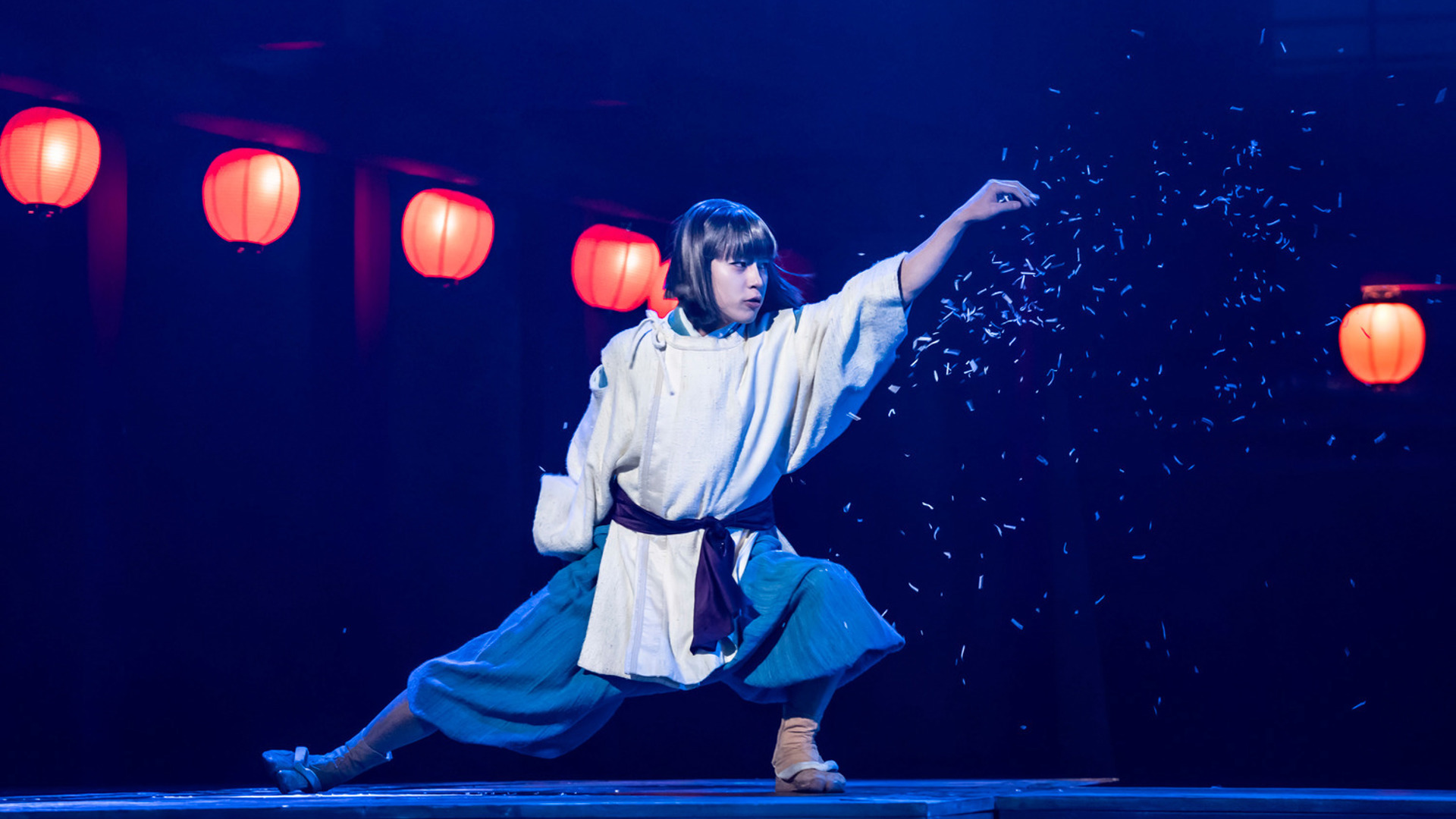 An actor stands in a sideways lunge, sprinkling confetti out of their left hand and looking to the left.