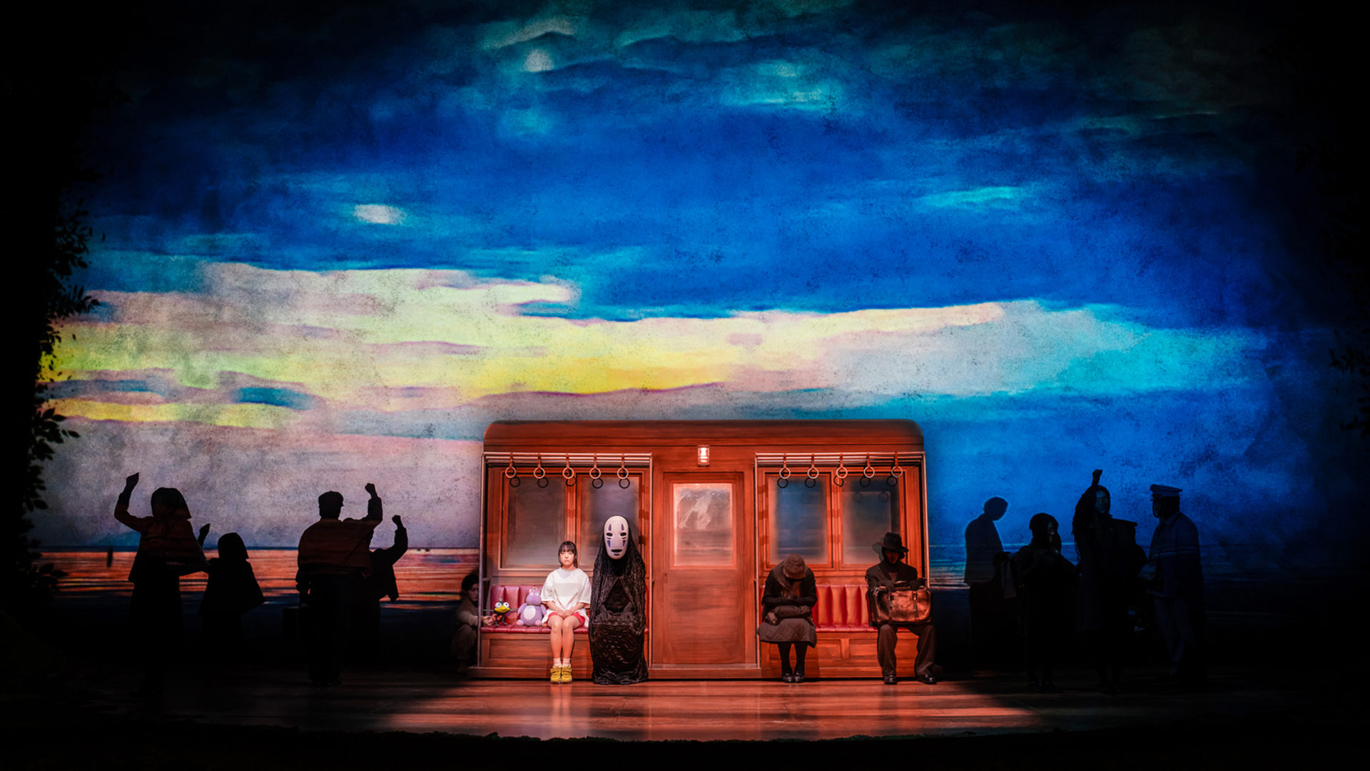 Two actors sit side by side on a train facing the audience. The actor on the right is wearing a white mask and black robes. The backdrop behind them is a mixture of blue and yellow sky.