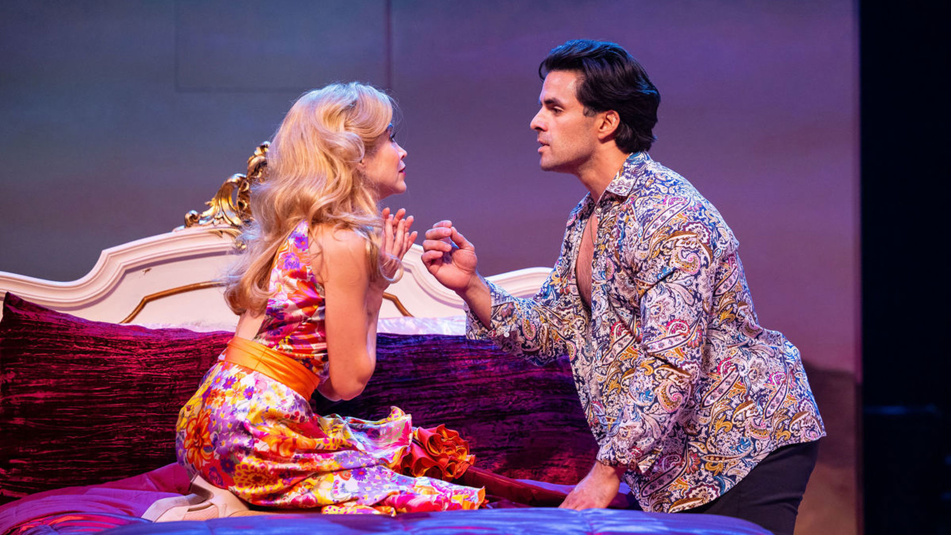 An actor kneels on a bed, holding their hands up, pleading to another actor who stands beside the bed.
