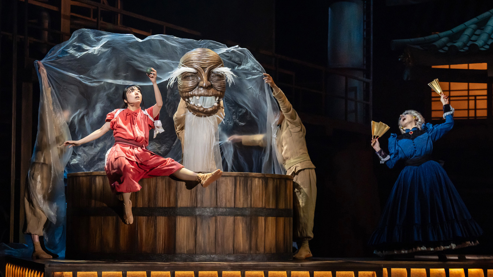 An actor dressed in all pink sits on the edge of a bath, looking at a small rock they are holding. There is a puppet of a large smiling face next to them. There is an actor to the right of the bath wearing a blue dress and holding two fans, looking feint.
