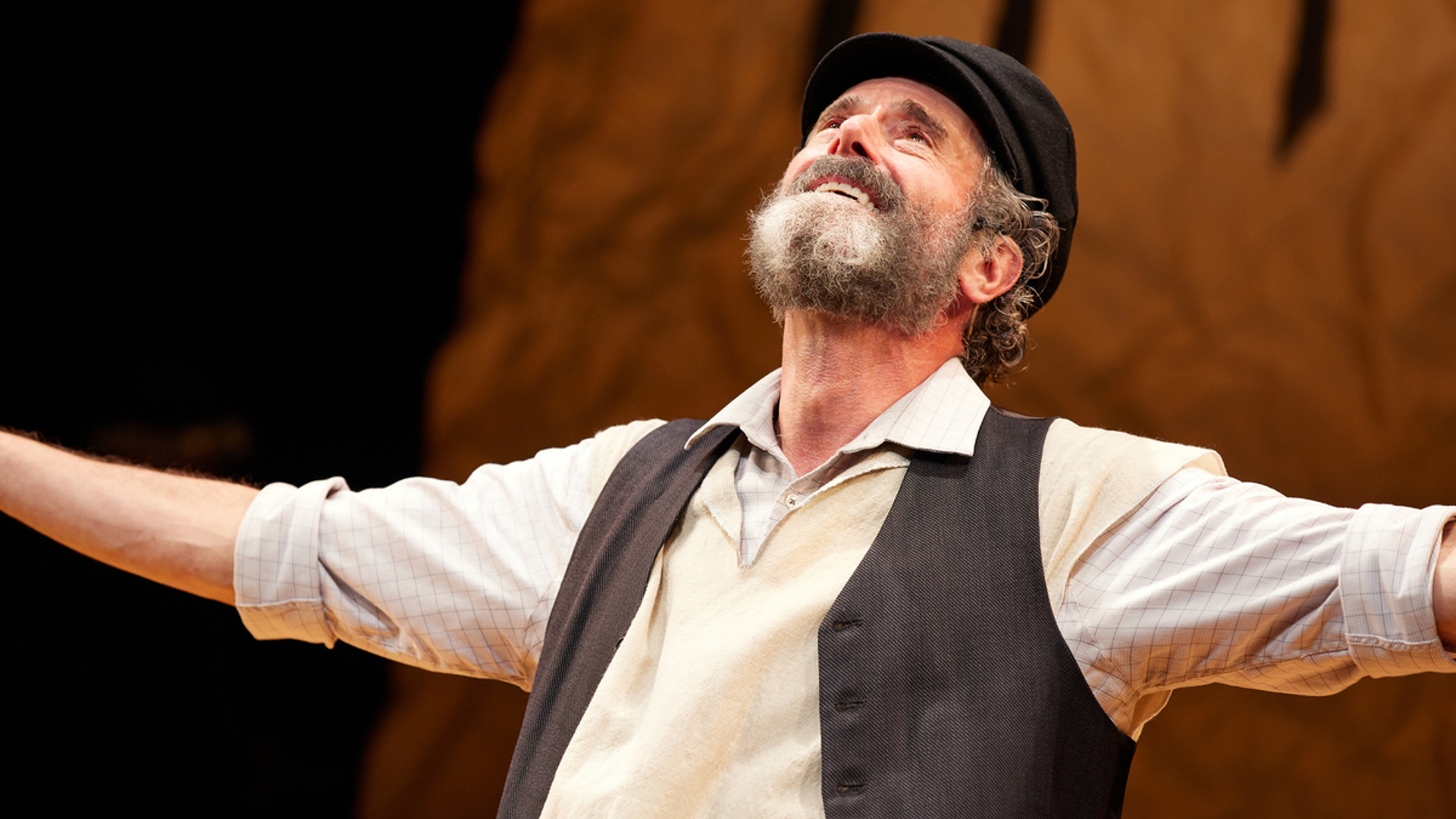 A man stands on stage stands with his arms out, he is smiling up at the sky. He is wearing a black hat and a gray vest as well as a tallit.