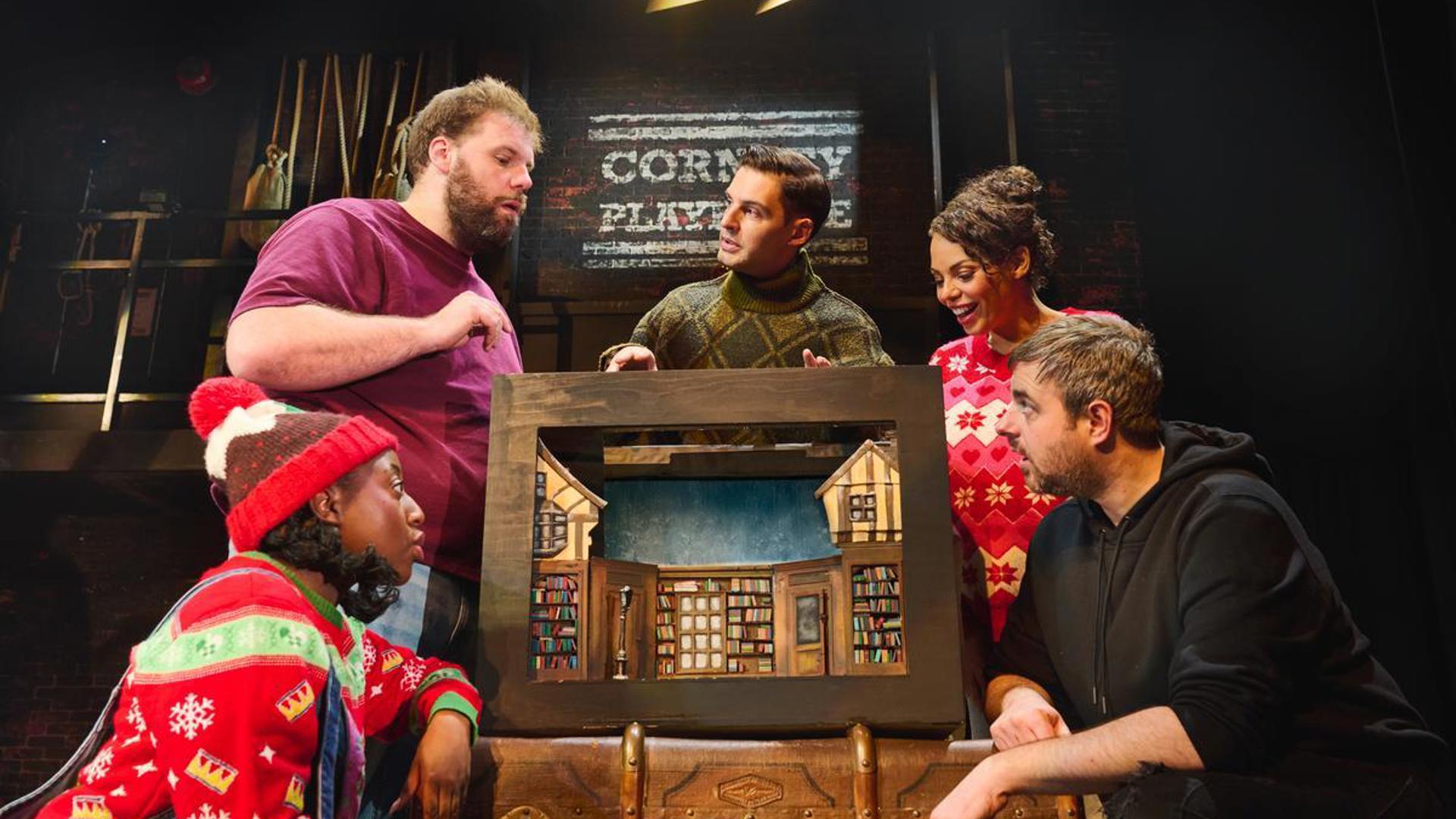 Five performers stand around a miniature set, looking in at the set.