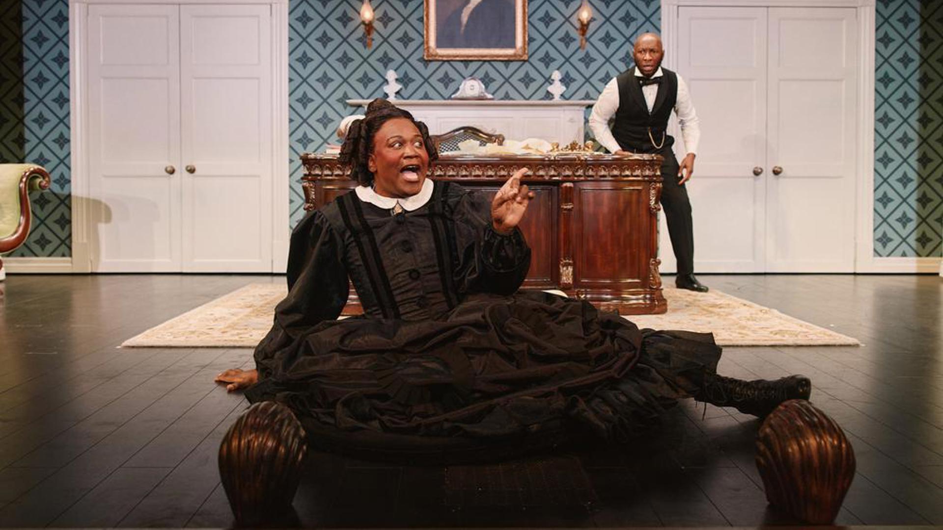 A performer sitting at the foot of the stage in front of a desk, another performer is coming around the desk behind them.
