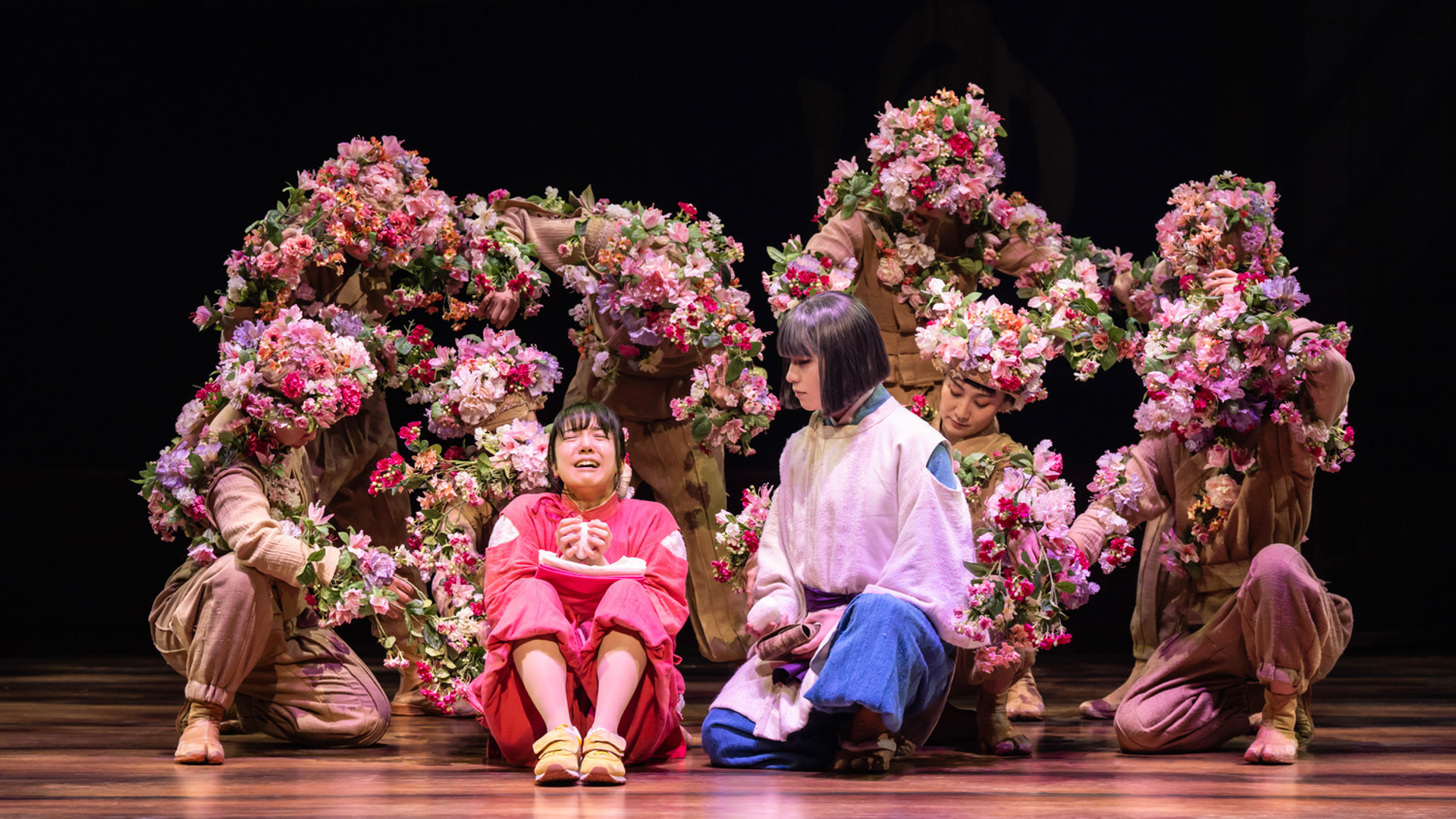 Two actors kneel side by side under a group of actors dressed as flower bushes. The actor on the left seems to be crying.