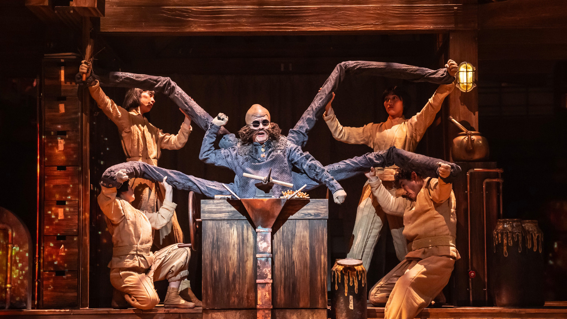 An actor with six arms controls a board of switches and levers. Other actors crowd around him, holding his arms.